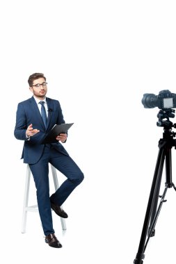 Beyaz, bulanık ön planda dijital kameranın yanındaki yüksek taburede oturan pano ile haber spikeri