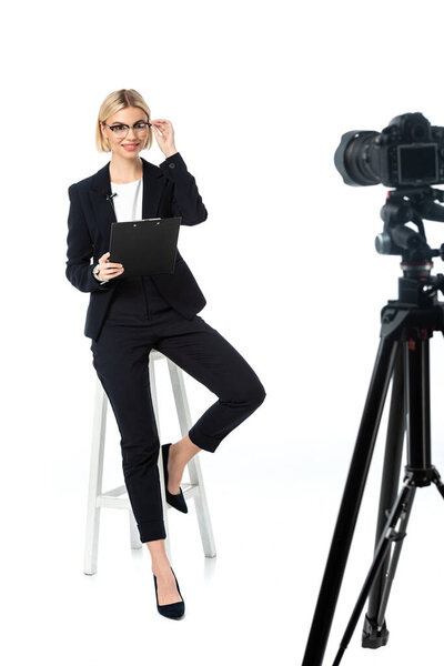 smiling news anchor touching eyeglasses while sitting on high stool near digital camera on white
