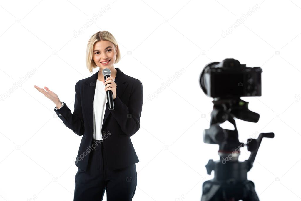 cheerful broadcaster with microphone pointing with hand near digital camera on blurred foreground isolated on white
