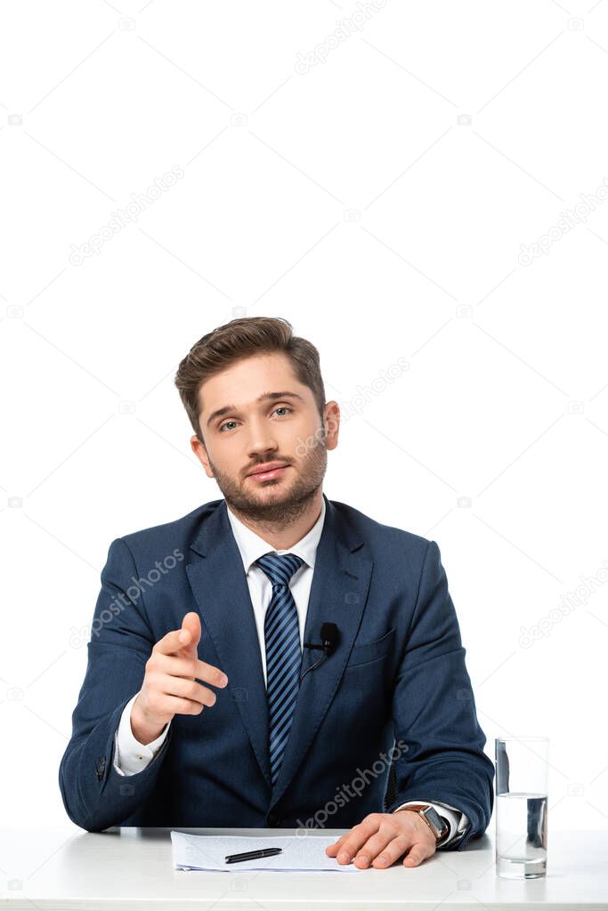 young news commentator pointing with finger near papers and glass of water isolated on white