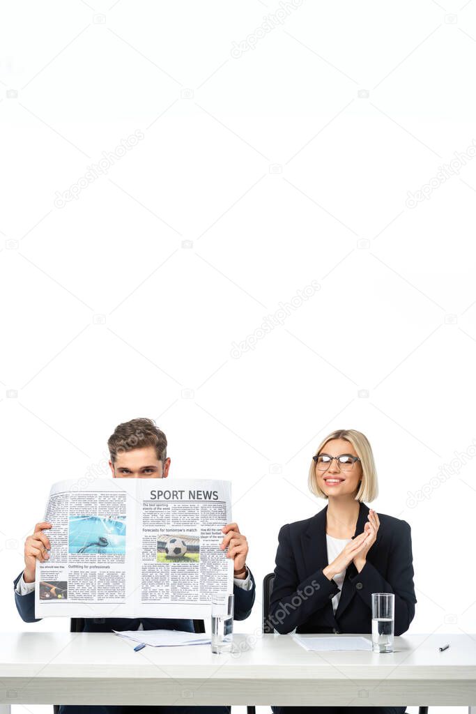 cheerful blonde journalist smiling near colleague obscuring face with newspaper isolated on white