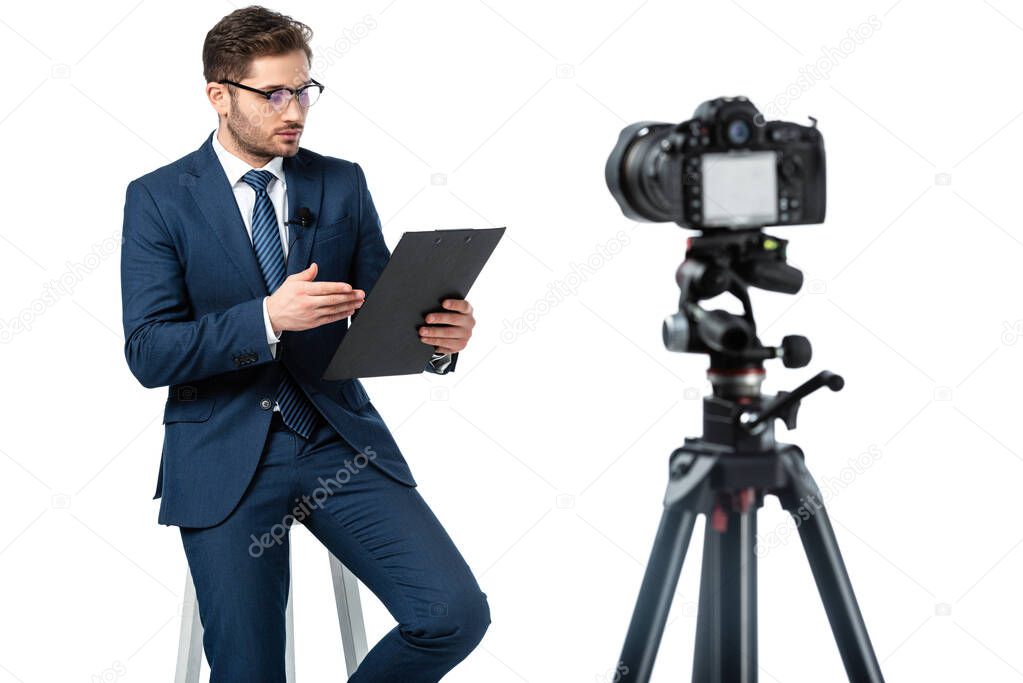 news anchor pointing at clipboard while sitting on high stool near digital camera on blurred foreground isolated on white