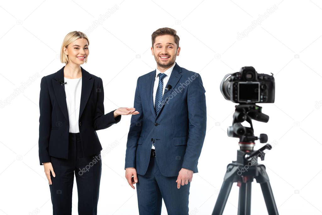 happy anchorwoman pointing at smiling colleague near digital camera on blurred foreground isolated on white