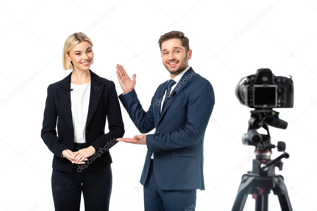 happy news anchor pointing at colleague near digital camera on blurred foreground isolated on white