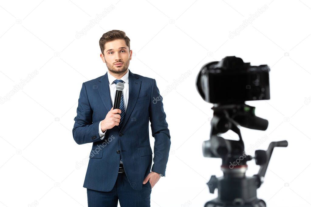 young broadcaster holding microphone near digital camera on blurred foreground isolated on white