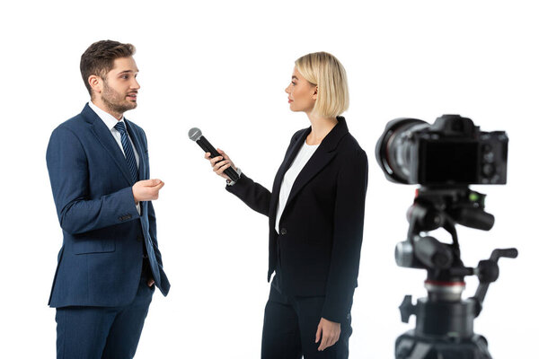 young businessman giving interview to blonde journalist with microphone near digital camera on blurred foreground isolated on white