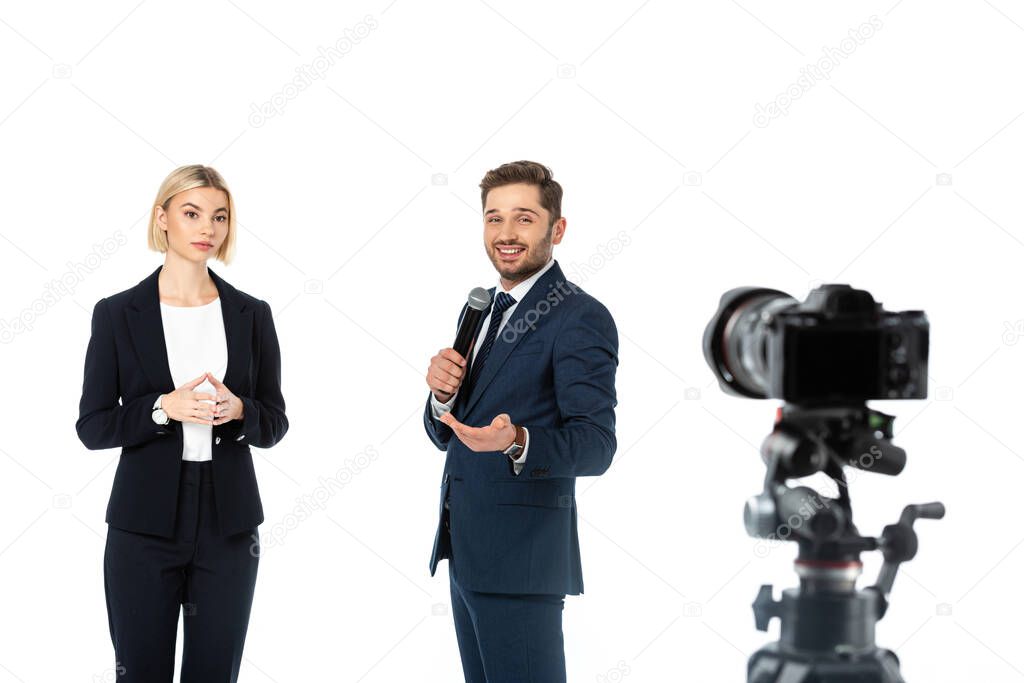 smiling broadcaster with microphone near blonde colleague and digital camera on blurred foreground isolated on white