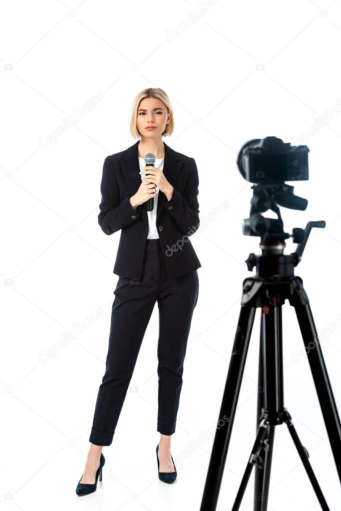 full length view of blonde news anchor with microphone near digital camera on white, blurred foreground