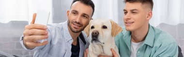 Aynı cinsiyetten neşeli bir çift gülümsüyor ve Labrador 'la selfie çekiyor.