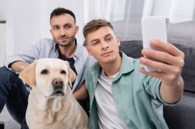 Aynı cinsiyetten çift, oturma odasındaki Labrador yakınlarında akıllı telefondan selfie çekiyor.