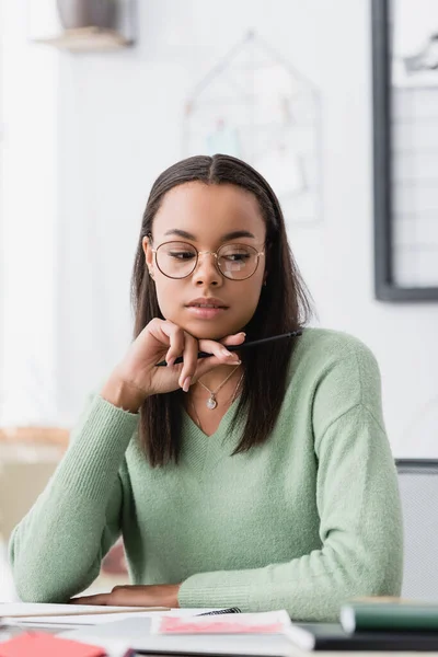 dreamy african american interior designer in eyeglasses holding pen while working at home
