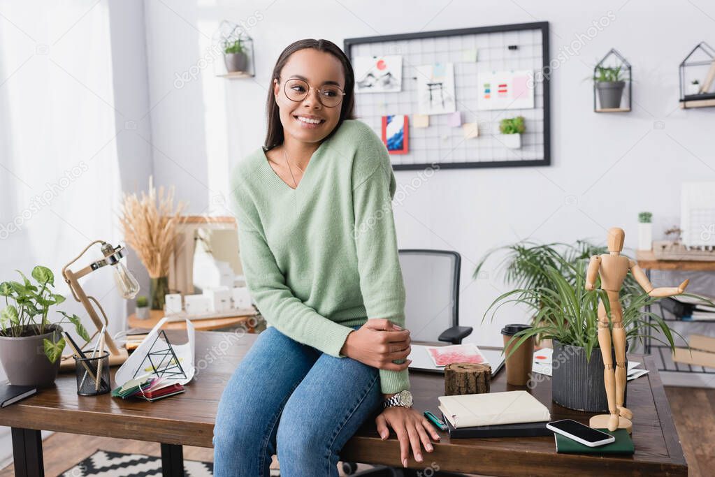 happy african american architect looking away while sitting on work desk at home