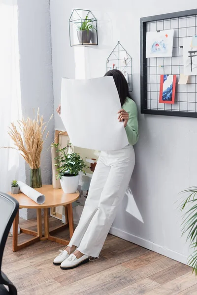 african american architect obscuring face with paper while leaning on wall at home