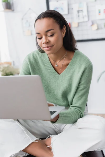 smiling african american interior designer using laptop while sitting with crossed legs at home