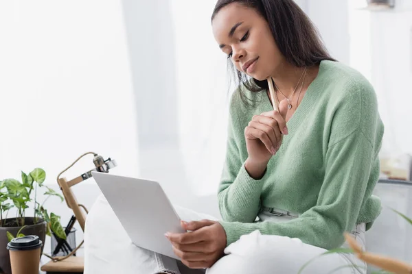 thoughtful african american interior designer holding pencil while sitting on desk with laptop
