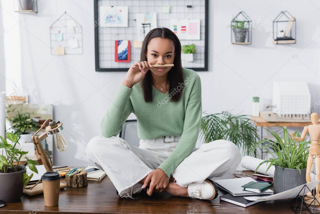 cheerful african american architect holding pencil near nose while sitting on desk with crossed legs