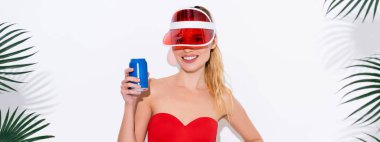 cheerful woman in sun visor smiling at camera while holding can of soda on white, banner clipart