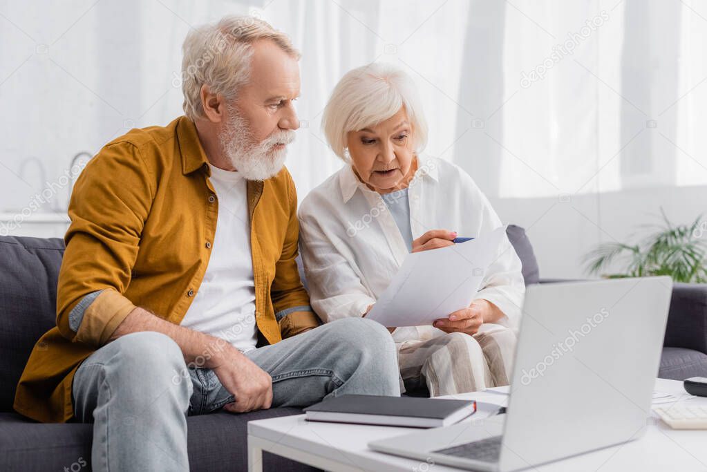 Senior woman holding papers near husband, notebook and laptop on blurred foreground 