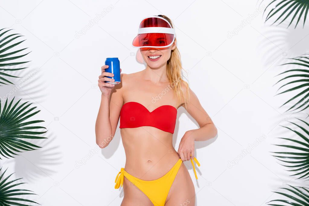 happy woman in sun visor and swimsuit holding soda while smiling at camera on white