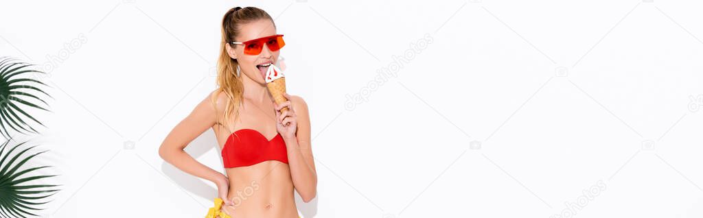 young woman in sunglasses and swimwear looking at camera while licking ice cream on white, banner