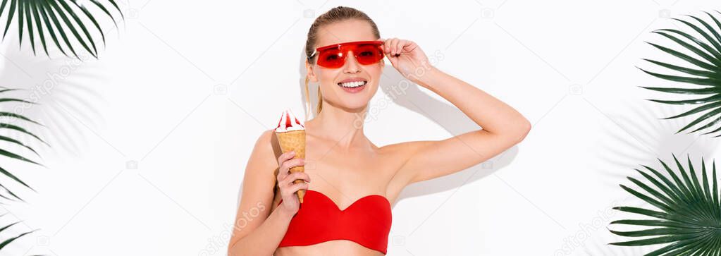 happy woman touching eyeglasses and smiling at camera while holding ice cream on white, banner
