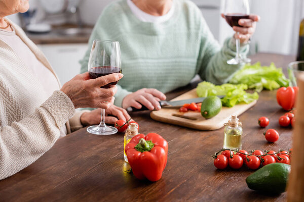 cropped view of senior women holding glasses of red wine near fresh vegetables in kitchen 