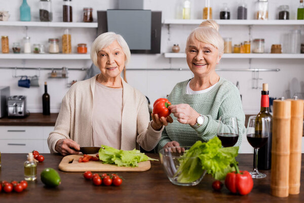 happy senior woman holding fresh bell pepper near retired friend in kitchen 