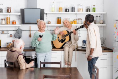 Mutfakta şarap kadehleriyle neşeli çok kültürlü arkadaşlarının yanında akustik gitar çalan son sınıf öğrencisi mutlu bir kadın. 