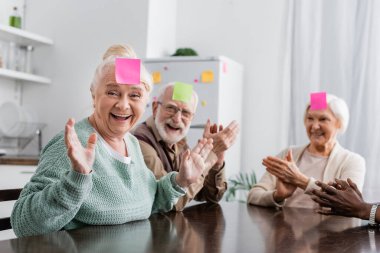 excited and multicultural senior friends with sticky notes on foreheads playing guess who game in kitchen  clipart