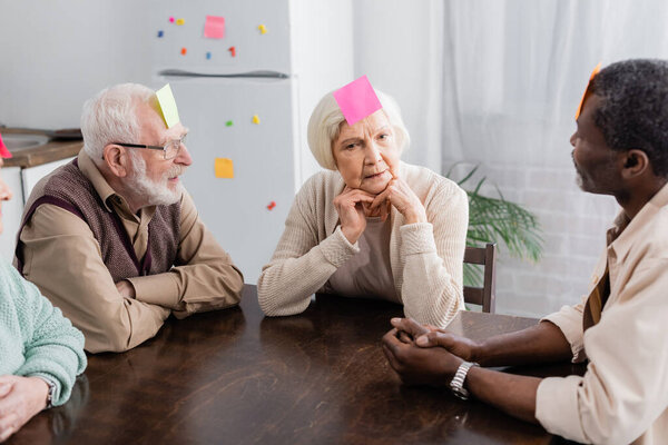 interracial senior friends with sticky notes on foreheads playing game in kitchen 