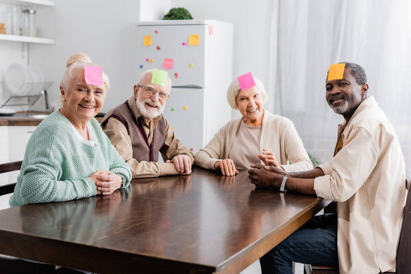 joyful multicultural senior friends with sticky notes on foreheads playing game in kitchen 