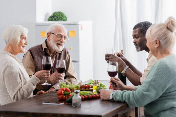 cheerful and retired interracial friends holding glasses with wine near vegetables on table