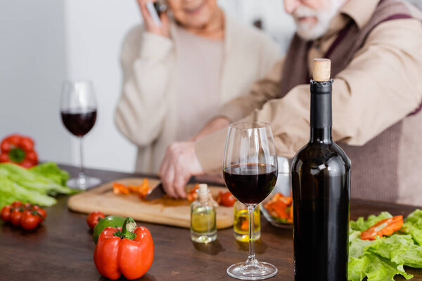 bottle with red wine near glass, vegetables and retired couple on blurred background 
