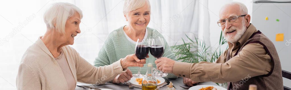 happy senior friends clinking glasses with red wine near tasty lunch on table, banner