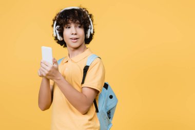 Sarıda izole edilmiş akıllı telefon kullanan kulaklıklı kıvırcık çocuk.