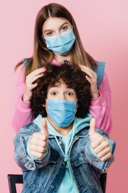 Koruyucu maskeli çocuk, bulanık ön planda, pembe bir arkadaşa yakın görünüyor. 