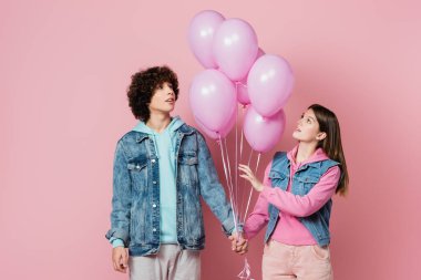 Gençler pembe arka plandaki balonlara bakarken el ele tutuşuyorlar. 