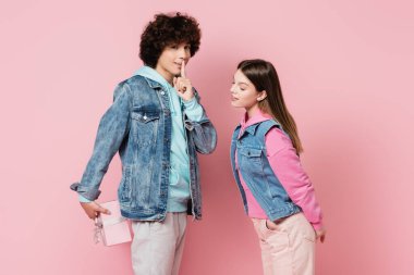 Teenager showing shh gesture while hiding present near girlfriend on pink background clipart