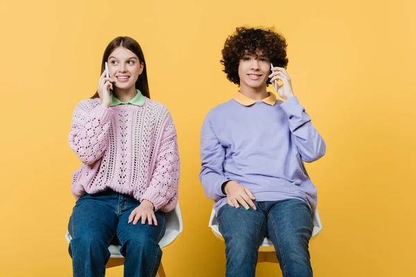 Uśmiechnięte Nastolatki Rozmawiające Przez Komórki Krzesłach Żółtym Tle — Zdjęcie stockowe
