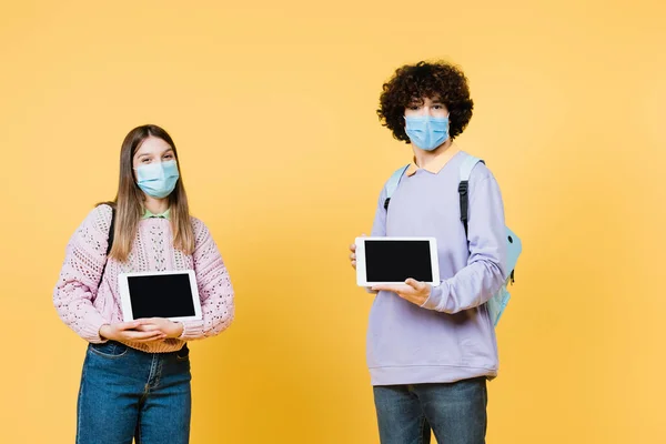 Nastolatki Maskach Medycznych Trzymające Tabletki Cyfrowe Białym Ekranem Żółtym Tle — Zdjęcie stockowe