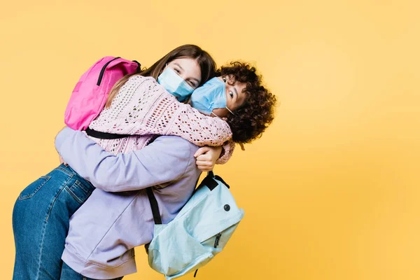 Adolescentes Con Máscaras Médicas Mochilas Abrazándose Aislados Amarillo — Foto de Stock