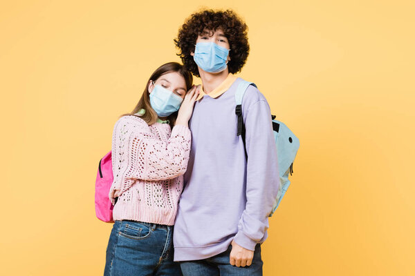 Teenage girl in medical mask embracing friend with backpack isolated on yellow