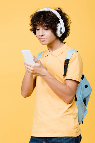 Teen boy listening music in headphones and using cellphone isolated on yellow