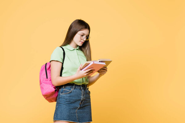 Girl in eyeglasses with backpack reading book isolated on yellow