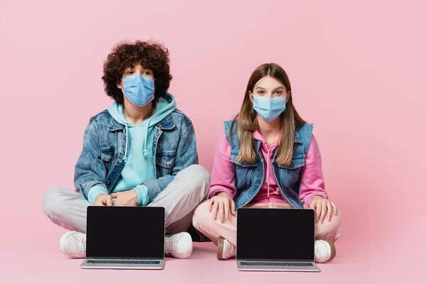 戴着口罩 双腿交叉坐在粉红背景笔记本电脑旁的青少年朋友 — 图库照片