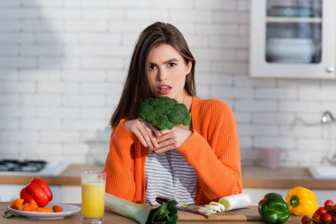 Ciddi bir kadın masada taze sebzelerin yanında brokoli tutarken kameraya bakıyor.