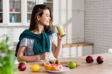 Mutfakta taze meyvelerin yanında bir bardak smoothie tutan neşeli kadın, bulanık ön plan.