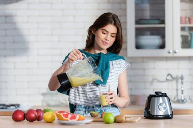Genç bir kadın mutfaktaki taze meyvelerin yanında blenderdan smoothie dolduruyor.