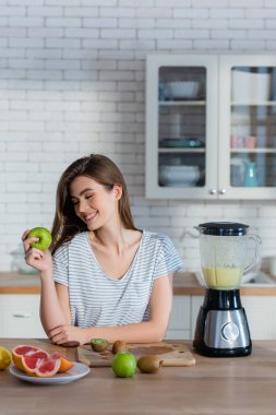 Mutfakta meyve ve blender yanında olgun elma tutan memnun bir kadın.