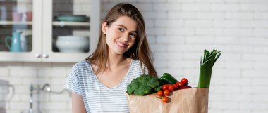 joyful woman looking at camera near fresh vegetables in paper bag, banner clipart